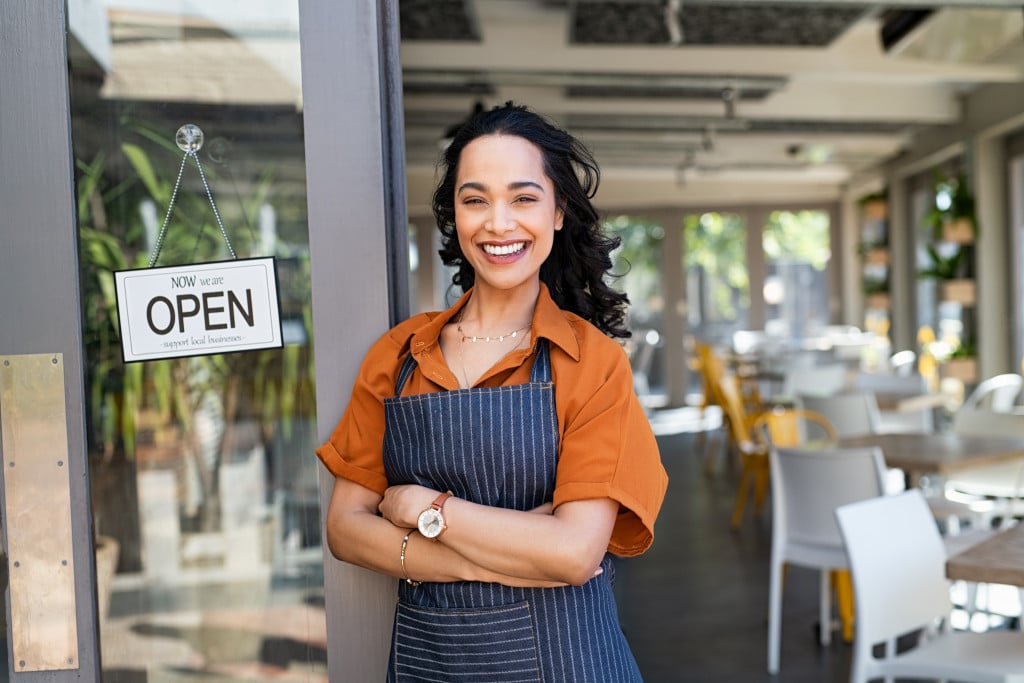 Happy female business owner standing in front of the door of a restaurant with an open sign.